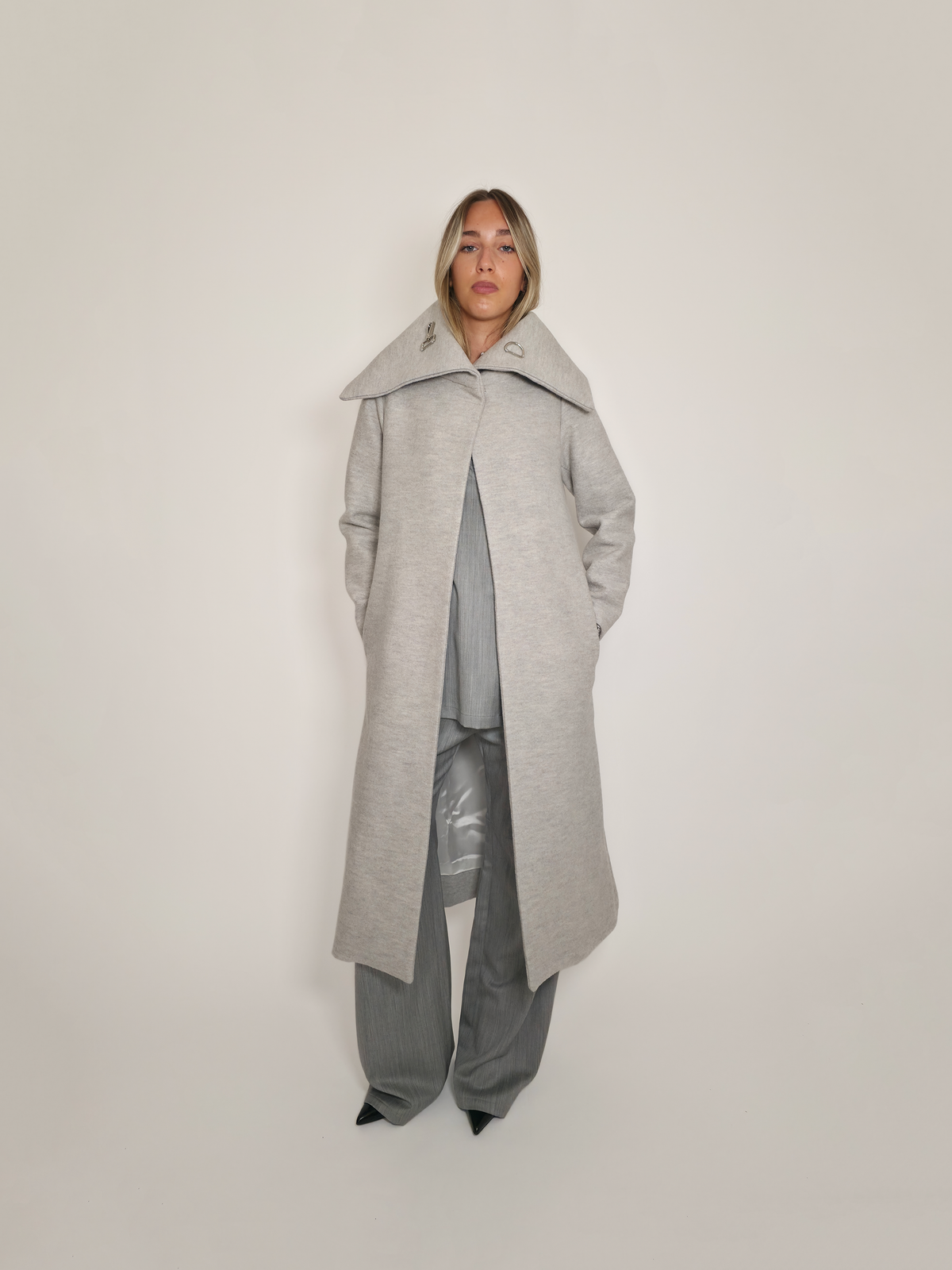 Femme portant le manteau long Michelle gris, fermé en haut, avec son grand col rabattu. Porté avec le pantalon Michael et la robe Myshellie.